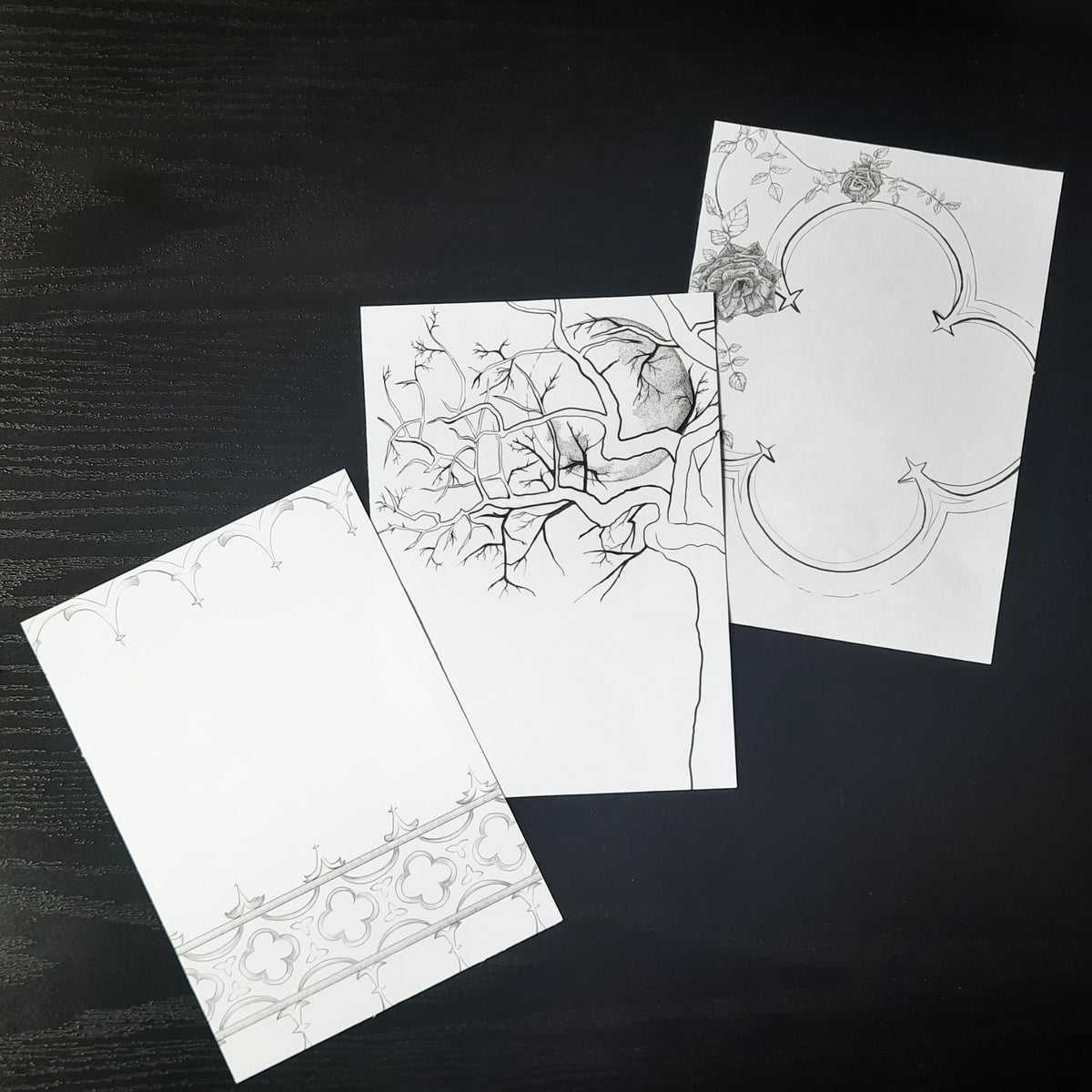 Three gothic themed black ink illustrations printed on white pages, laying on a dark background.