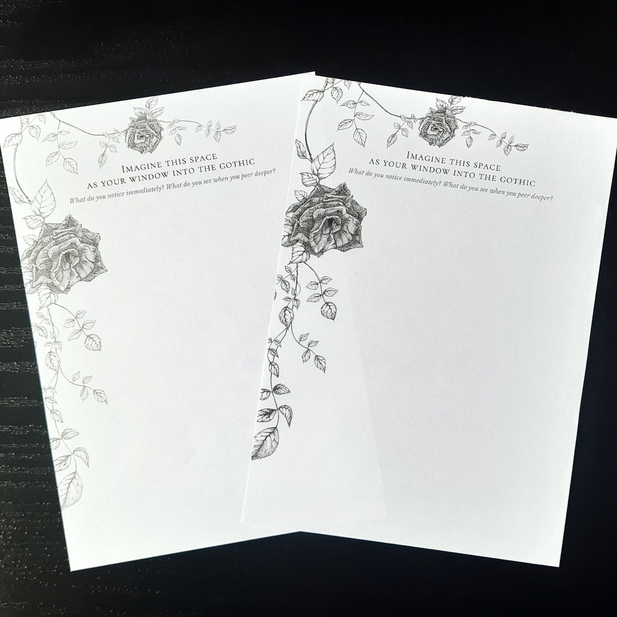 Two gothic themed black ink illustrations of roses with a written journal prompt, printed on white pages, laying on a dark background.