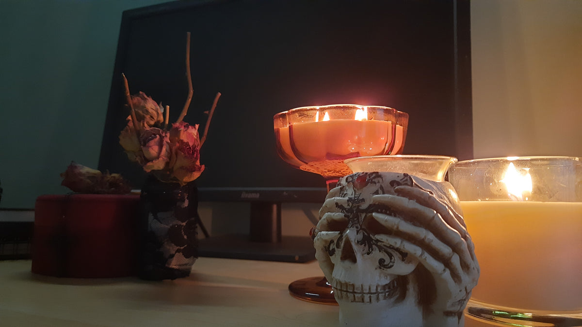 A gothic-themed tabletop arrangement featuring a ceramic skull holding a candle, a lit amber glass goblet candle, a lit white candle in a glass holder, and dried flowers in a dark vase against a dark monitor background.